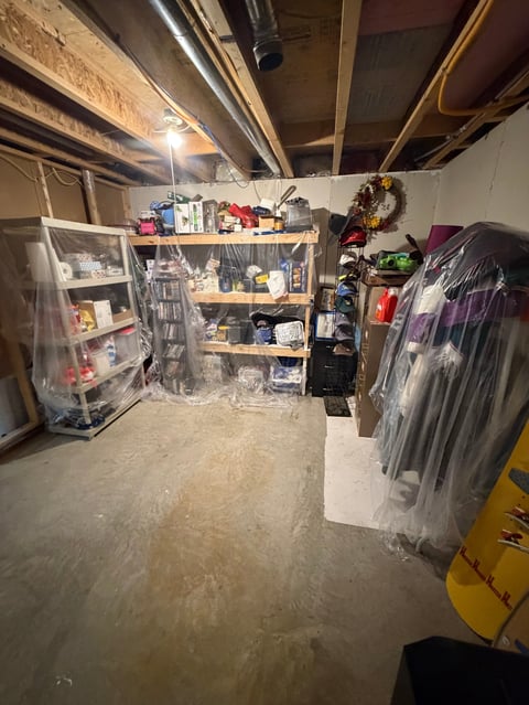 Cluttered basement storage area with wooden shelving, plastic storage bins, hanging clothes, and various boxes under exposed ceiling beams and lighting.