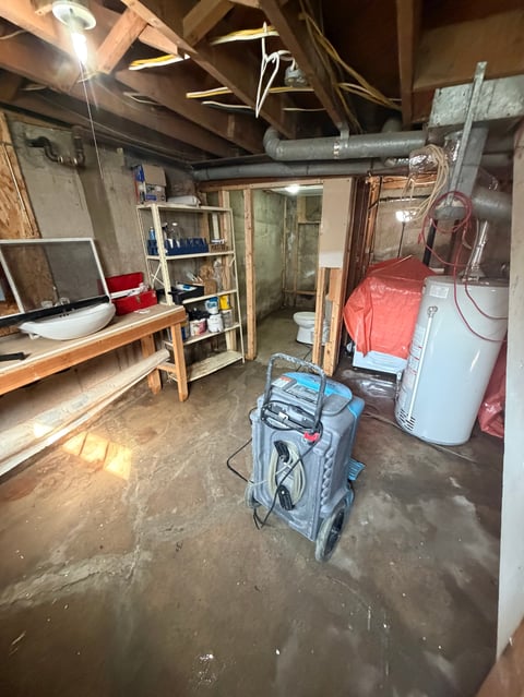 Unfinished basement with wooden workbench, shelving, dehumidifier, water heater, and various tools and equipment on concrete floor.